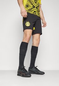 Svarta shorts med en gul klubb-logotyp och två gula sidostråk, ihopparade med svarta knä-långa strumpor och svarta träningsskor med röda detaljer.