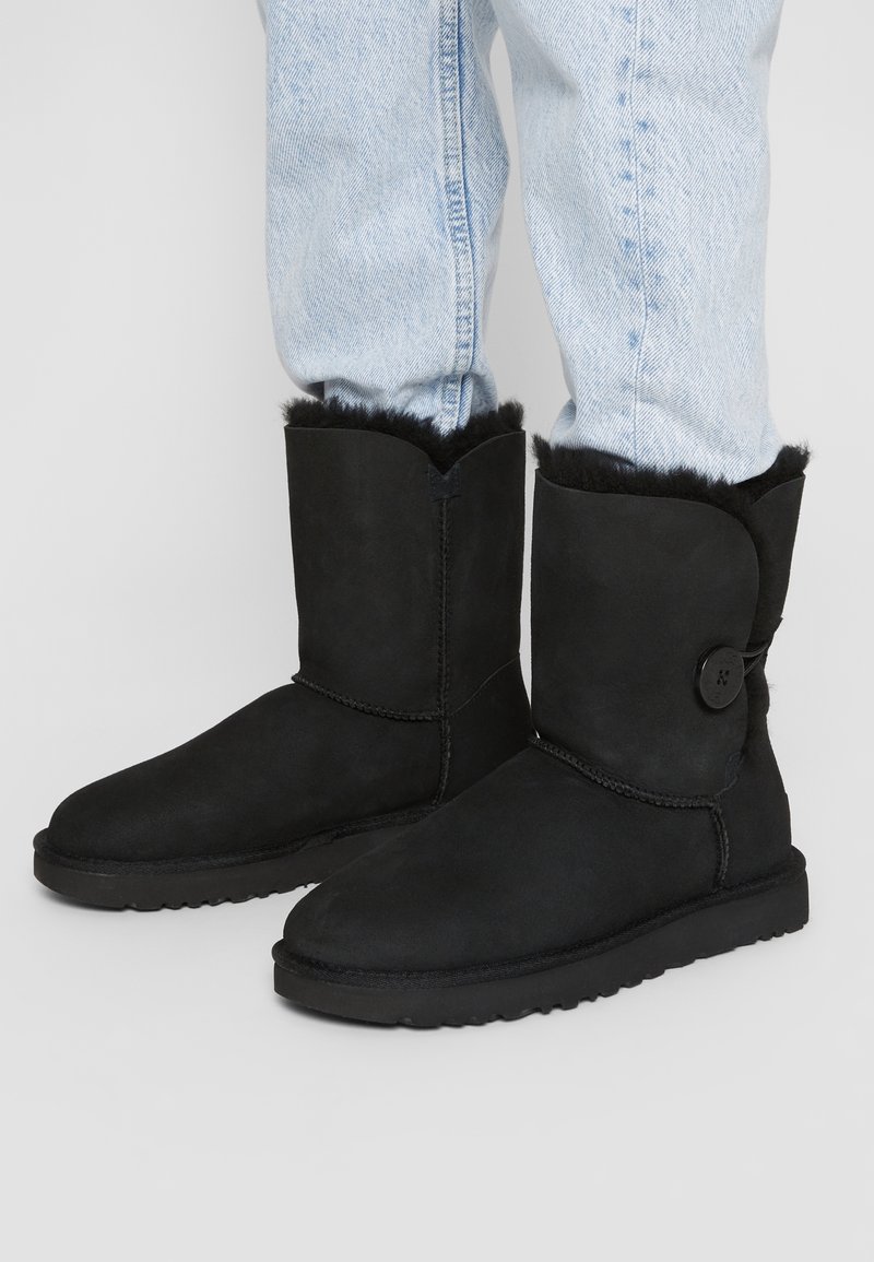 Personne portant un jean bleu clair et des bottes mi-mollet en suède noir avec une doublure en fourrure et un bouton sur le côté extérieur de chaque botte.