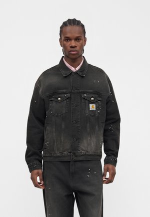 Junger Mann mit geflochtenem Haar, der eine schwarze Jeansjacke und passende Hose mit weißen Farbflecken vor einfarbigem Hintergrund trägt.