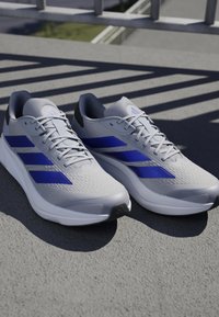 Gray athletic shoes with blue accents, textured mesh upper, and padded collar. Features prominent three-stripe logo and cushioned sole.