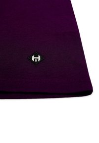 Lilla bomulds t-shirt med rund halsudskæring, korte ærmer og et sort diamantformet logo-patch med et hvidt bogstav "R" nær kanten.