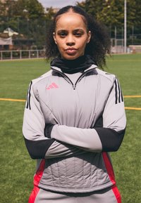 Mujer con cabello rizado y pecas que lleva una chaqueta deportiva negra y gris con acentos rojos, de pie con los brazos cruzados en un campo de fútbol.