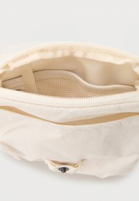 Μπεζ υφασμάτινη θήκη με ανοιχτό φερμουάρ, που εμφανίζει εσωτερική τσέπη από δίχτυ και κούμπωμα με κουμπί στο μπροστινό καπάκι.