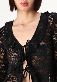 Top in pizzo nero caratterizzato da motivi floreali, scollatura a scallop e chiusura a lacci frontale, con una texture leggera e traslucida.