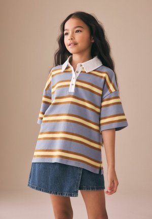 Jeune fille aux longs cheveux portant un polo rayé jaune, marron et lavande, ainsi qu'une jupe en jean, sur un fond beige.