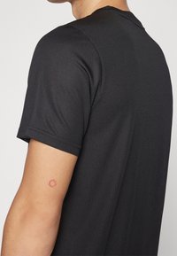 Person som bär en randig svart kortärmad tröja, visar en liten röd cirkulär tatuering på överarmen nära ärmen.