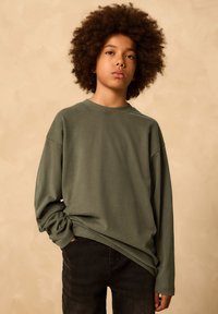 Tânăr cu păr creț, purtând o cămașă cu mâneci lungi de culoare verde măsliniu și pantaloni negri, pe un fundal simplu bej.