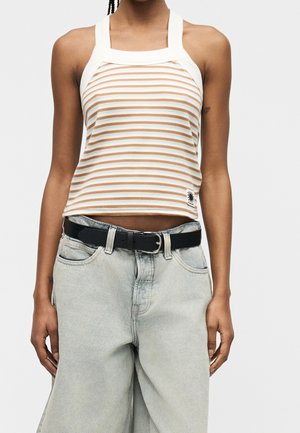Kvinde iført en beige og hvid stribet haltertop med lysegrå højtaljede jeans og et sort bælte, står foran en ensfarvet baggrund.