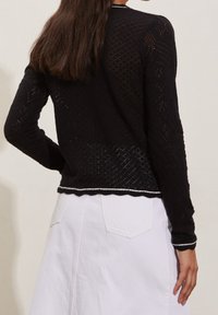 Pull en maille noire avec un motif texturé, manches longues et ourlet festonné avec des accents blancs ; associé à une jupe blanche.