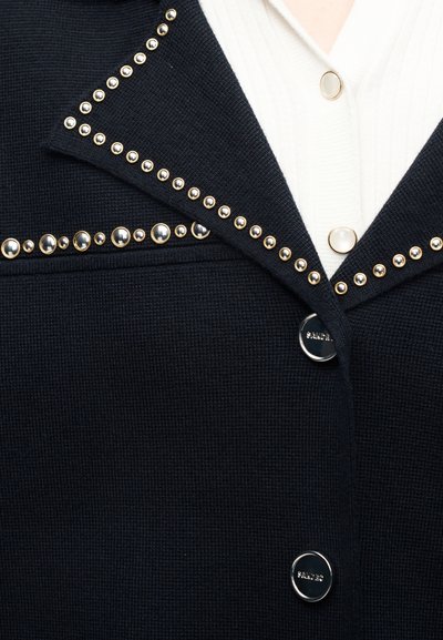 Μαύρο υφασμάτινο μπουφάν με λαμπερές λεπτομέρειες διακοσμημένες με χρυσά καρφιά στον γιακά, φορεμένο πάνω από ένα λευκό πουλόβερ με κουμπιά που μοιάζουν με πέρλες.