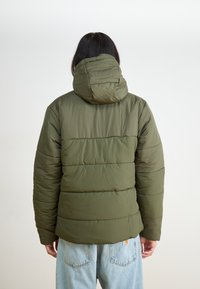 Tamsiai žalia pūkinė striukė su gobtuvu, su įsiūtais raštais ir horizontaliomis siūlėmis. Dėvima su šviesiai mėlynomis džinsinėmis kelnėmis.