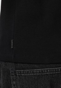 Schwarzes, geripptes Stoffoberteil mit einem kleinen Markenschild nahe dem Saum, kombiniert mit dunklem Denim. Die Textur ist glatt mit deutlichen vertikalen Linien.