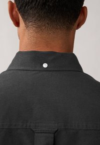 Maglietta nera con colletto a punta, con un singolo bottone sul retro e un tessuto testurizzato. Il retro presenta un design minimalista senza motivi.