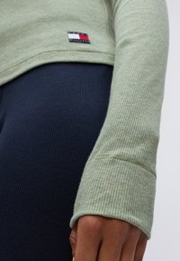 Lichtgroene geribbelde langemouwen top met een prominente merklogo patch; close-up van de mouw toont textuur en pasvorm tegen een marineblauwe achtergrond.