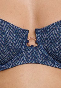 Top bikini blu scuro con motivo a zigzag, dotato di un accento metallico circolare al centro e tessuto testurizzato. Spalline regolabili.