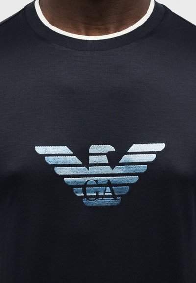 Μπλε ναυτικό T-shirt με άσπρο ριμπ λαιμόκομμα; διαθέτει μεγάλο, υφασμάτινο λογότυπο με διαβαθμισμένα χρώματα σε αποχρώσεις του μπλε, που απεικονίζει στυλιζαρισμένα φτερά και γράμματα.