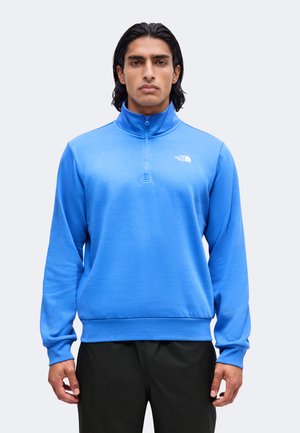 Homme aux cheveux foncés portant un sweat bleu à fermeture éclair en quart avec le logo The North Face, debout devant un fond clair uni.