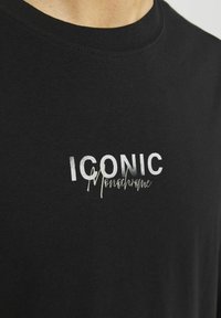 Czarna bawełniana koszulka z okrągłym dekoltem. Zawiera wyraz "ICONIC" napisany dużą białą czcionką oraz "Monochrome" w kursywie. Gładka tekstura.