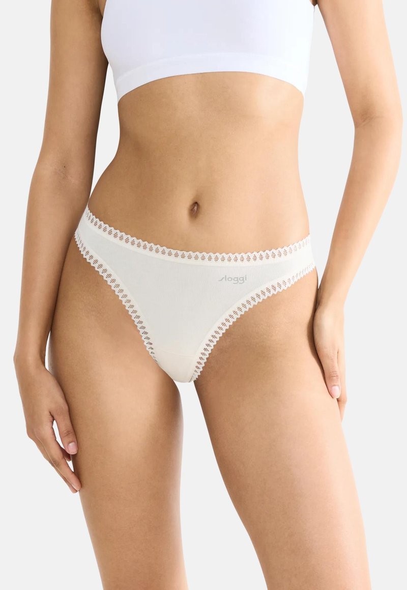 Sous-vêtements crème avec une bordure en dentelle festonnée, présentant une texture lisse et un design taille basse. Logo de la marque visible à l'avant.