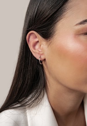 Nahaufnahme vom linken Ohr einer Frau mit silbernem Creolen-Ohrring und glattem dunklem Haar, die ein weißes Kleidungsstück trägt.