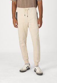 Beige Jogginghosen mit elastischem Bund und Kordelzug. Sie verfügen über schwarze Reißverschlusstaschen und schmal zulaufende Bündchen und sind aus einem weichen Material gefertigt.