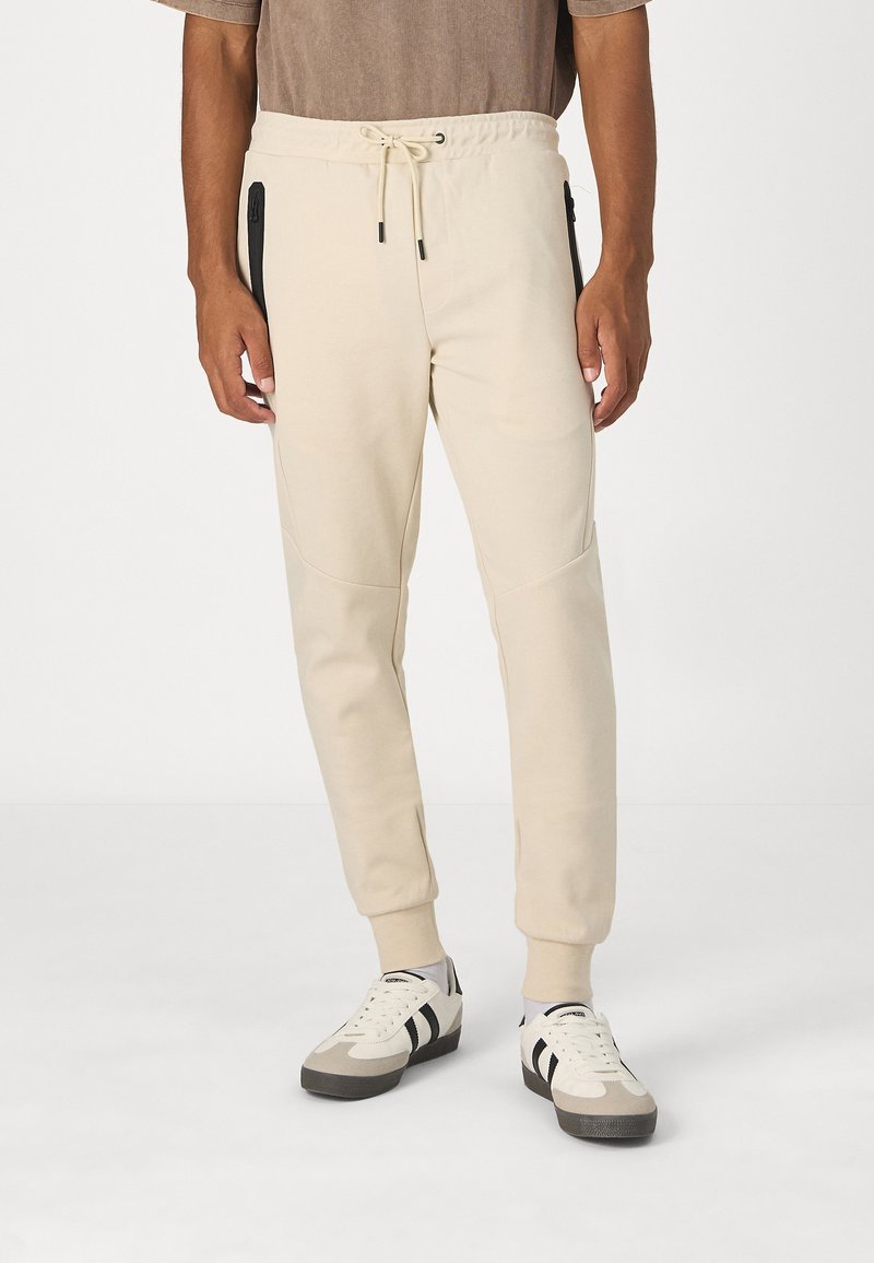 Beige Jogginghosen mit elastischem Bund und Kordelzug. Sie verfügen über schwarze Reißverschlusstaschen und schmal zulaufende Bündchen und sind aus einem weichen Material gefertigt.
