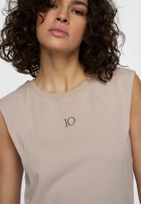 Top maniche corte di colore beige chiaro in un tessuto morbido, con collo rotondo e dettaglio ricamato "10" in marrone scuro centrato sul davanti.