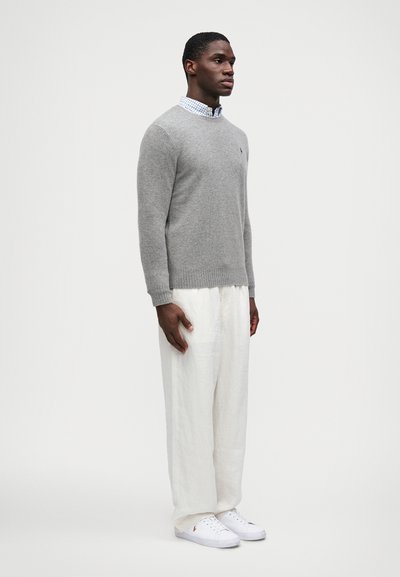 Pulôver cinza de malha com decote redondo, combinado com calças de linho brancas, largas e leves, e ténis brancos. Detalhe discreto do logótipo no pulôver.