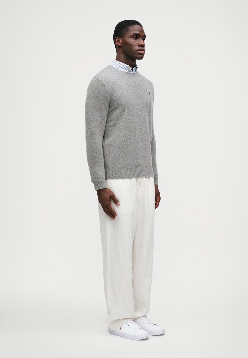 Suéter gris de punto con cuello redondo, combinado con pantalones de lino blanco, ligeros y holgados, y zapatillas blancas. Detalle de logo sutil en el suéter.