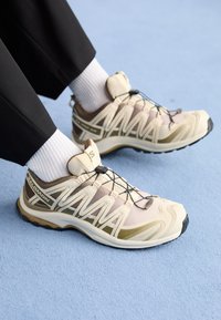 Jasne beżowe buty sportowe z siateczkową i syntetyczną cholewką, z brązowym akcentem, elastycznymi sznurówkami oraz teksturowaną gumową podeszwą.