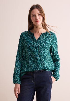 Blusa verde con motivo a foglia, scollatura a V e dettagli arricciati al collo. Maniche lunghe con polsini elasticizzati, abbinata a jeans scuri.