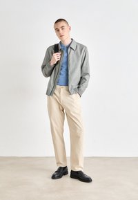 Svetlo sivá kockovaná bomber bunda so zipsom vpredu; modré polo tričko pod ňou; béžové nohavice; čierne slip-on topánky. Minimalistický dizajn.