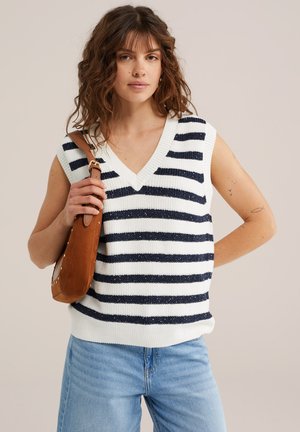 Donna con capelli castani mossi, che indossa un maglione smanicato a righe bianche e blu navy, jeans blu chiaro e tiene una borsa a tracolla marrone.