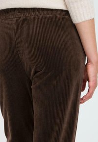 Brown corduroy pants feature vertical ribbing, elastic waistband, and a smooth texture. The focus is on the rear fit and seam details.