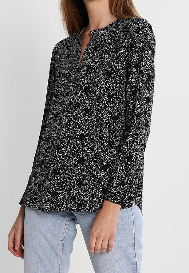 Femme portant un chemisier noir à manches longues avec de petits pois blancs et des motifs floraux, associé à un jean bleu clair.