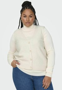 Elfenbensfärgad stickad väst med ett texturerat mönster, med v-ringning och fyra knappstängningar, lagd över en vit polotröja och bärs med blå jeans.