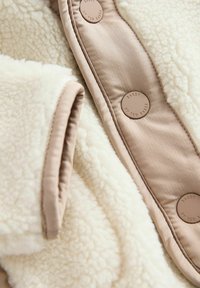 Pruunikas-kreemjas fliisjakk pehme tekstuuriga, millel on ümmargused pronksist nupud ja siledad pruuni kanga detailid ääreli.