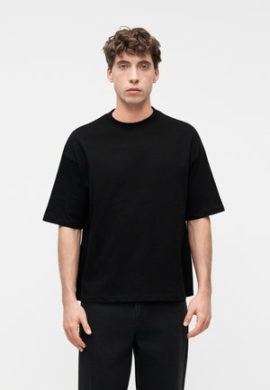 Fiatal férfi göndör barna hajjal, sima fekete, túlméretezett rövidujjú ingben és fekete nadrágban, fehér háttér előtt állva.