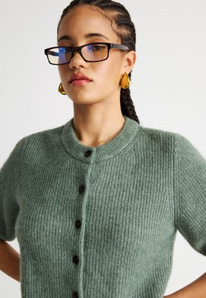 Γυναίκα με πλεξούδες, που φορεί γυαλιά με μαύρο σκελετό, χρυσούς σταγονένιους σκουλαρίκια και πράσινο πλεκτό πουλόβερ με κουμπιά, κοιτάζοντας ελαφρώς προς τα κάτω.