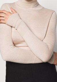 Person som bär en figursydd beigefärgad polotröja och korsar armarna över bröstet, i kombination med en svart kjol eller byxor.