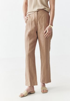 Pantaloni di lino marrone chiaro con design a gamba dritta e tasche laterali, abbinati a sandali neutri e una maglietta beige con texture.