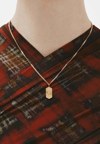 Collar con colgante de oro en forma rectangular que presenta un diseño de estallido solar en una fina cadena, llevado sobre un textil oscuro de color rojo con patrones.