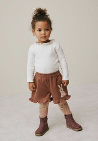 Lille pige med krøllet hår iført hvid langærmet bluse, brune flæse shorts med kanin-applikation, stribede strømpebukser og brune støvler, der står indendørs.
