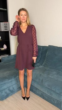 Vestido bordô com decote em V e cintura com laço, com mangas transparentes e pontilhadas. Combinado com saltos pretos de bico fino. Fundo com sofá azul texturizado.