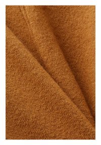 Gros plan sur un tissu en laine camel douce et texturée avec un motif tricoté visible et de légers plis.