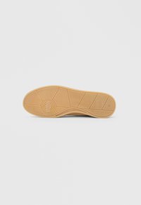 Bėgio padas, pagamintas iš kreminės spalvos tekstūruoto guminio paviršiaus su zigzaginių ir apskritimų sukibimo raštais ant balto fono.