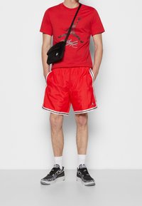 Czerwona koszulka z krótkim rękawem z graficznym wzorem, połączona z błyszczącymi czerwonymi szortami z białym wykończeniem i logo. Czarne sneakersy z wzorzystymi akcentami.