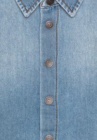 Helles blaues Jeanshemd mit einem Kragen und sechs Metallknöpfen auf der Vorderseite, sichtbaren Nähten und einer weichen, strukturierten Oberfläche.