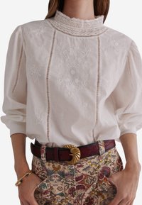 Blusa bianca ricamata con colletto alto in pizzo e maniche a sbuffo. Presenta dettagli a taglio lungo le cuciture verticali. Abbinata a pantaloni con motivo.