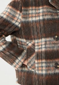 Veste en laine à carreaux aux nuances de marron, crème et orange, avec une texture douce, de gros boutons et une poche latérale.